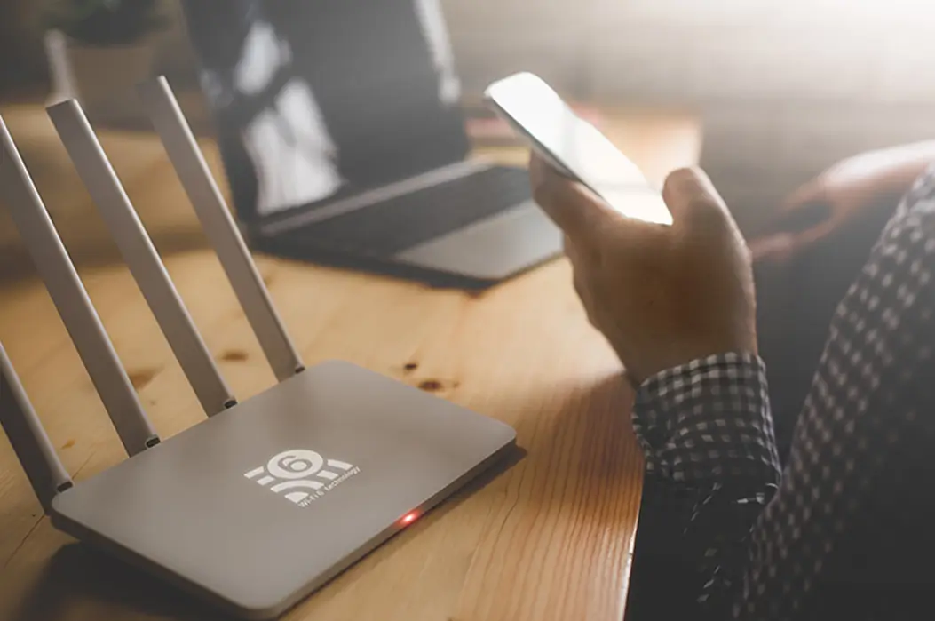 Ein moderner WLAN-Router auf einem Holzschreibtisch, daneben eine Person mit Smartphone