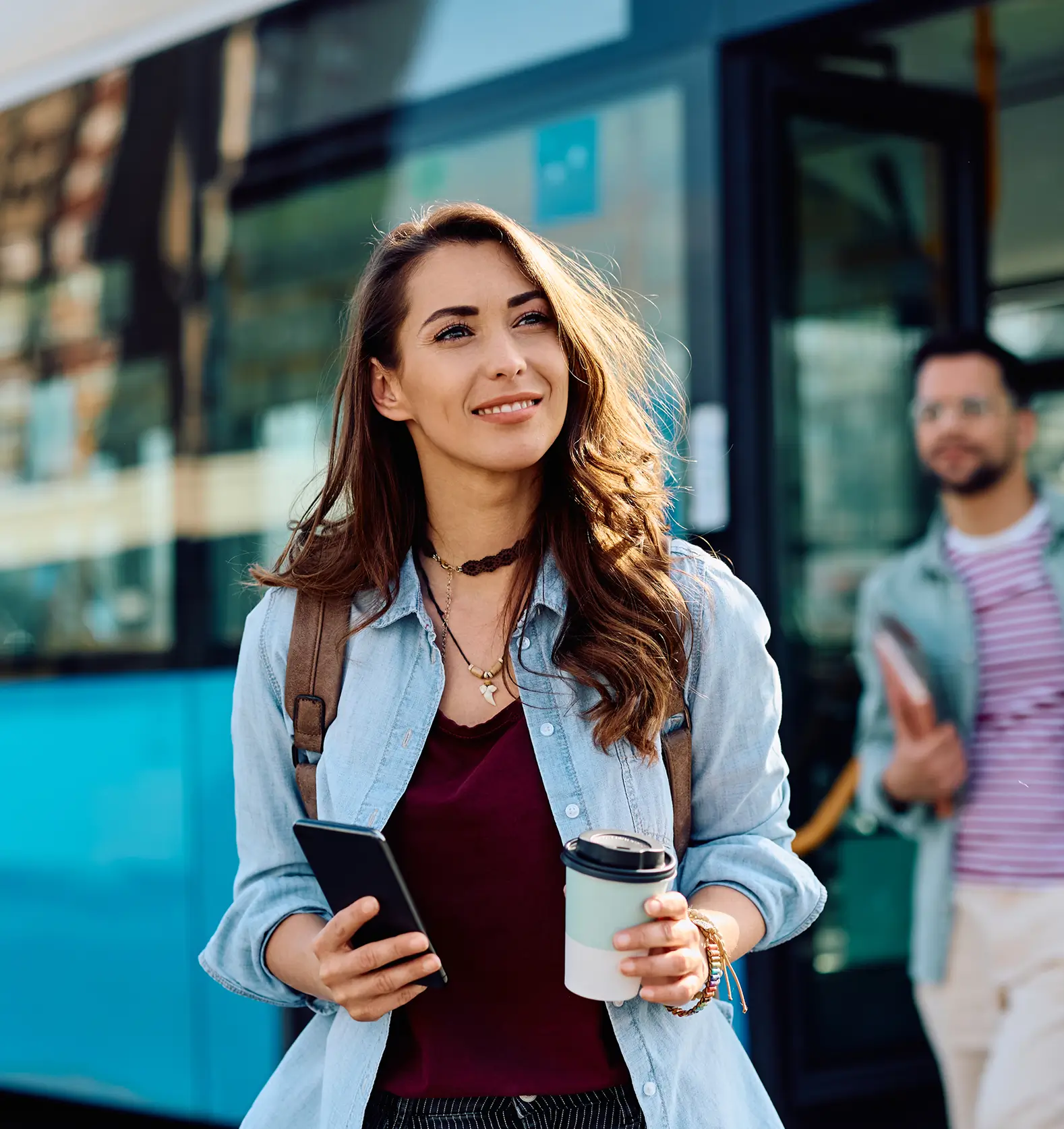 Frau steht mit Smartphone und Kaffee vor einem Bus, Mann im Hintergrund