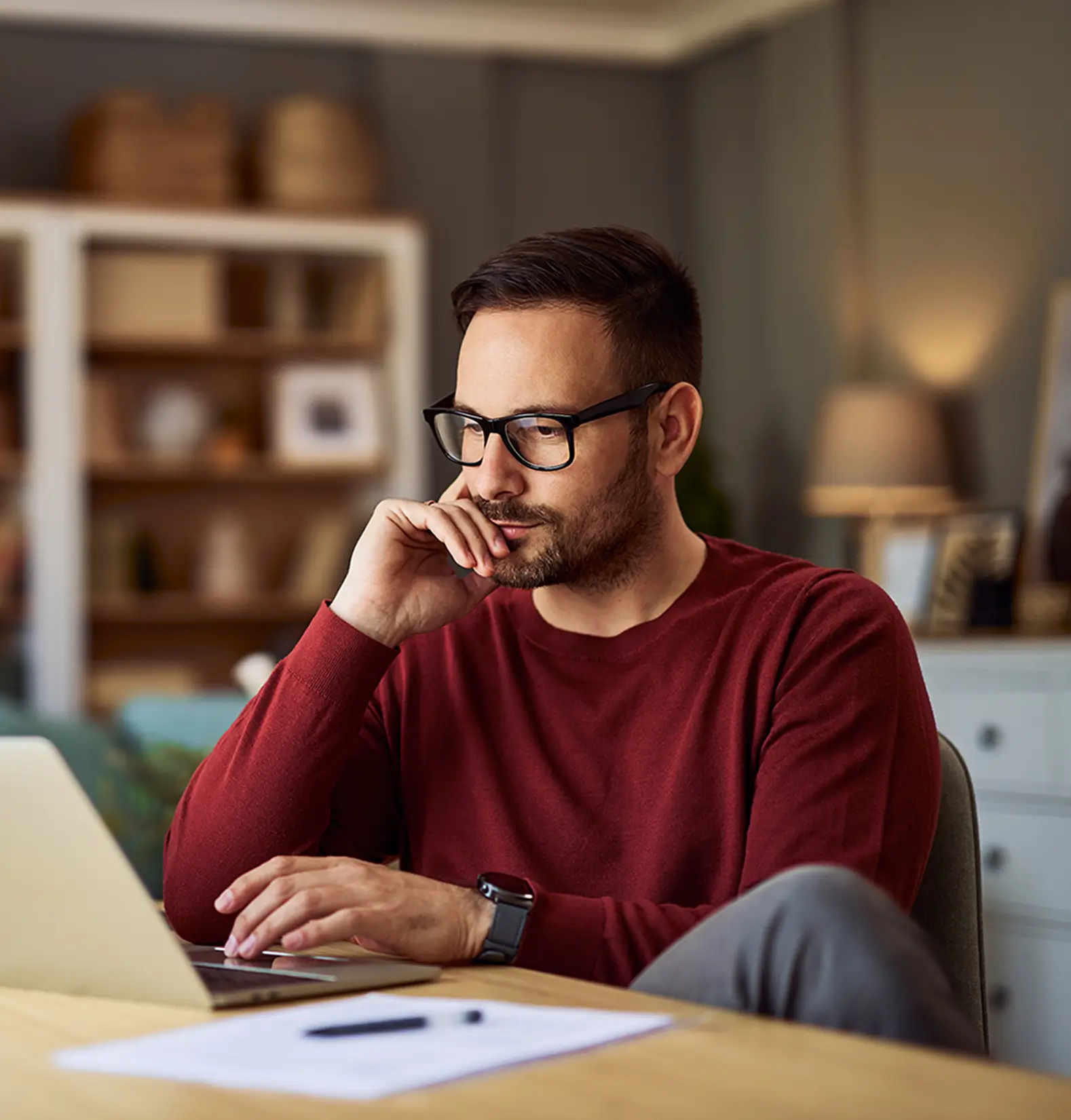 Mann mit Brille und rotem Pullover sitzt vor Laptop und denkt nach