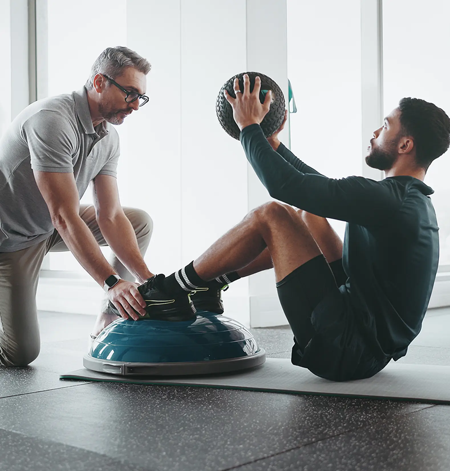 Trainer assistiert einem Sportler beim Training mit einem Medizinball auf einer Halbkugel