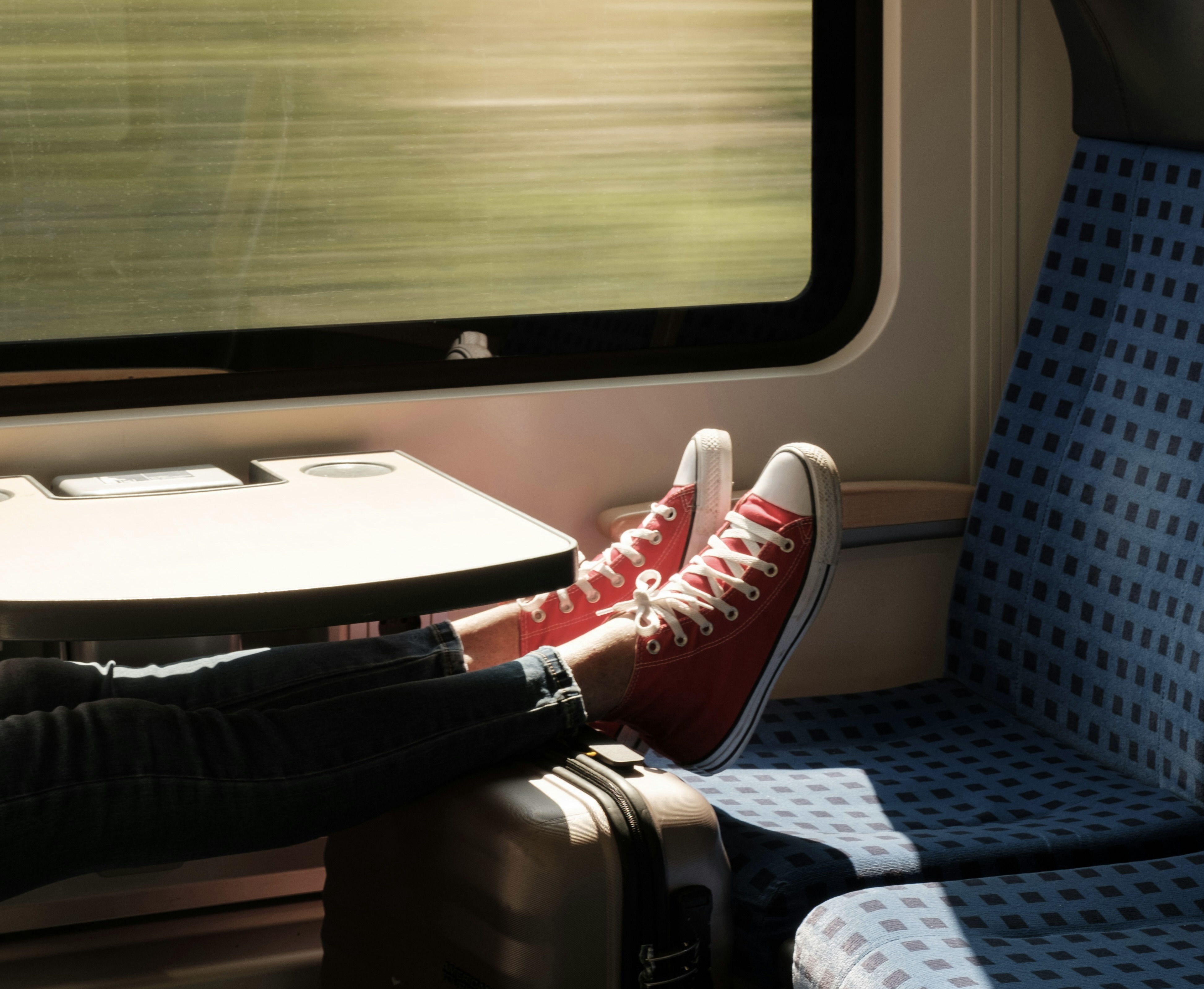 Eine Person in einem Zugabteil lehnt entspannt die Füße in roten Sneakern auf einen Koffer, während draußen die Landschaft vorbeizieht.